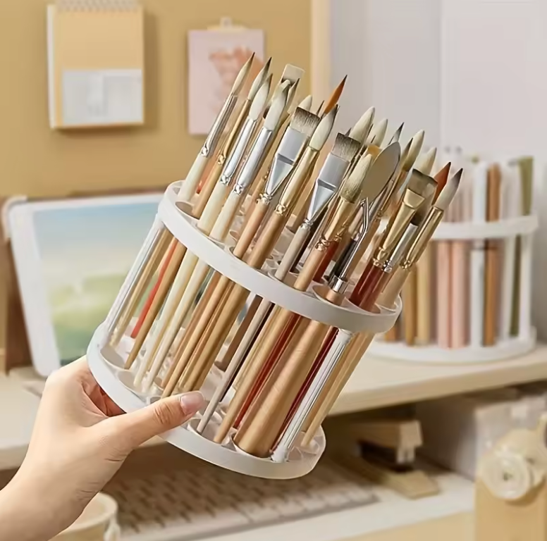 Cylindrical 360° rotating brush holder filled with art brushes, held by a hand in a workspace with art supplies.