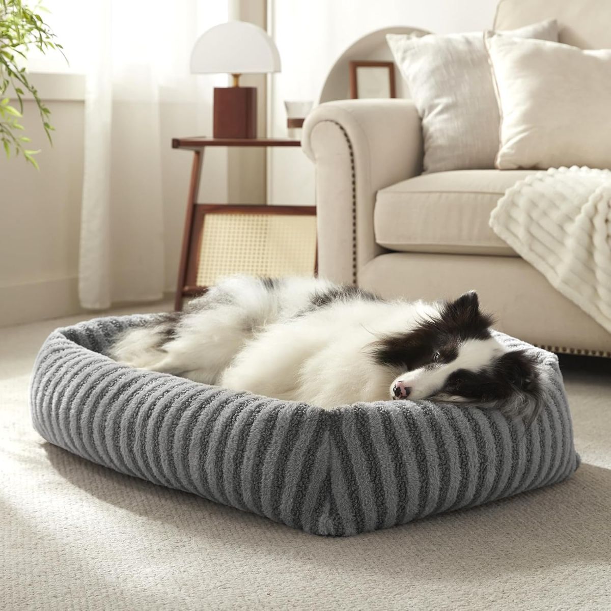 Striped gray dog bed with raised edges, placed on a light-colored floor, surrounded by soft furnishings.
